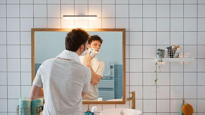 Mann rasiert sich vor einem Badezimmerspiegel, der mit einer Philips LED-Wandlampe beleuchtet wird.