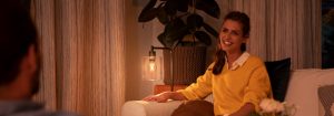 „Gemütliche Wohnzimmeratmosphäre durch eine warme LED-Tischlampe, die eine Frau im gelben Pullover bei einem abendlichen Gespräch beleuchtet.“