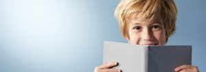 "Jeune enfant aux cheveux blonds souriant derrière un livre ouvert, présentant une expérience de lecture joyeuse sur fond bleu clair. »